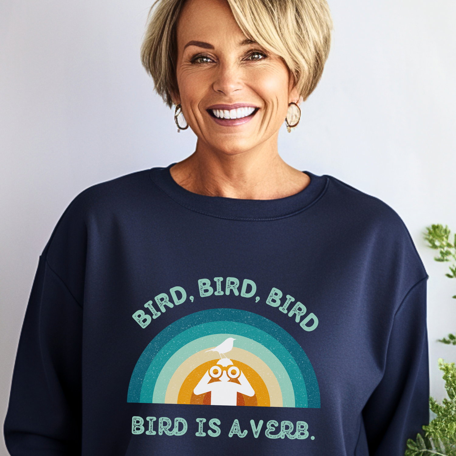 Smiling woman wearing a navy blue sweatshirt featuring a playful and colorful design of a rainbow and a bird perched on a figure holding binoculars. The text reads, 'BIRD, BIRD, BIRD. BIRD IS A VERB.'